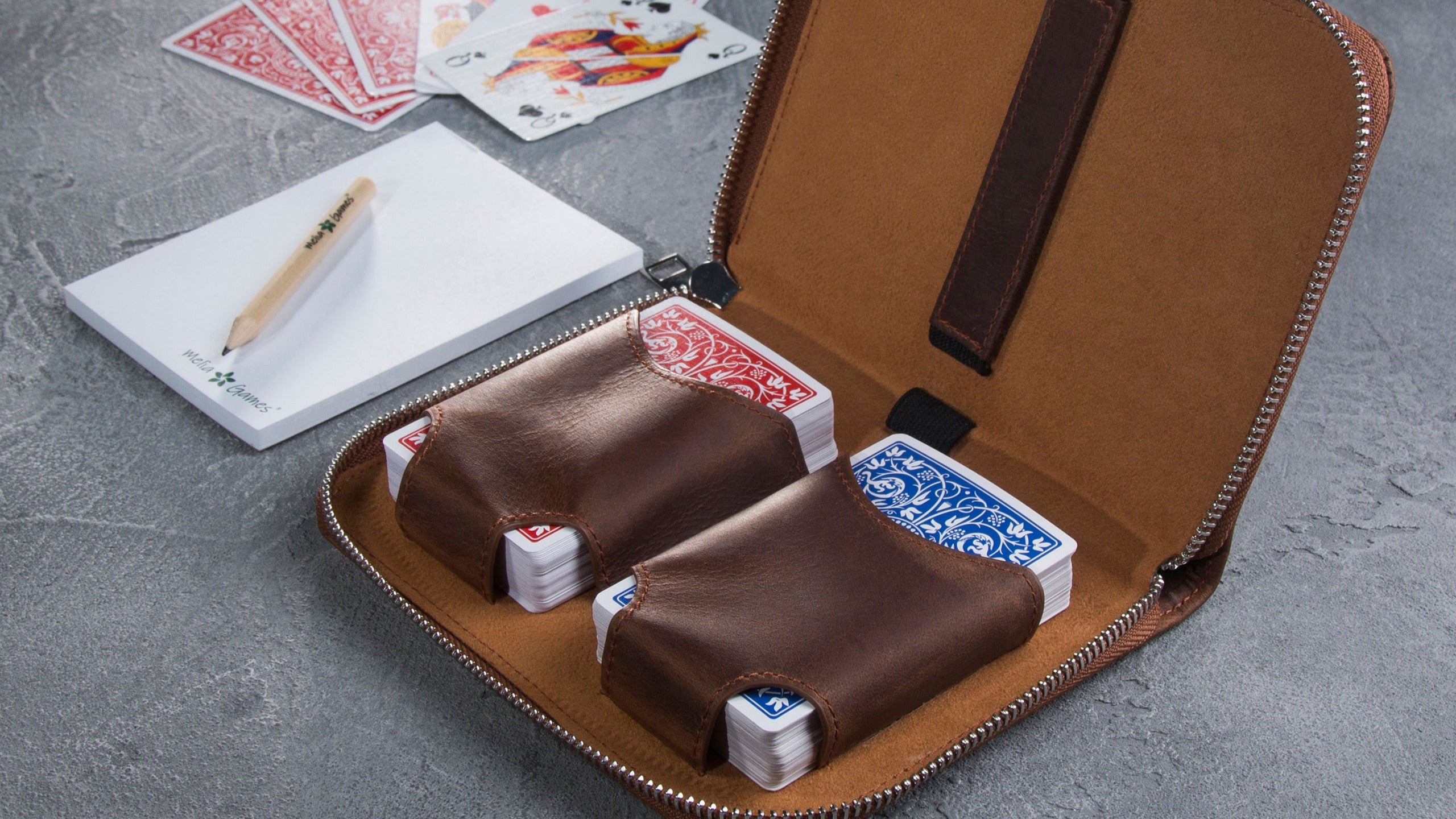 Brown leather case with playing cards, pen, and notebook on a gray surface